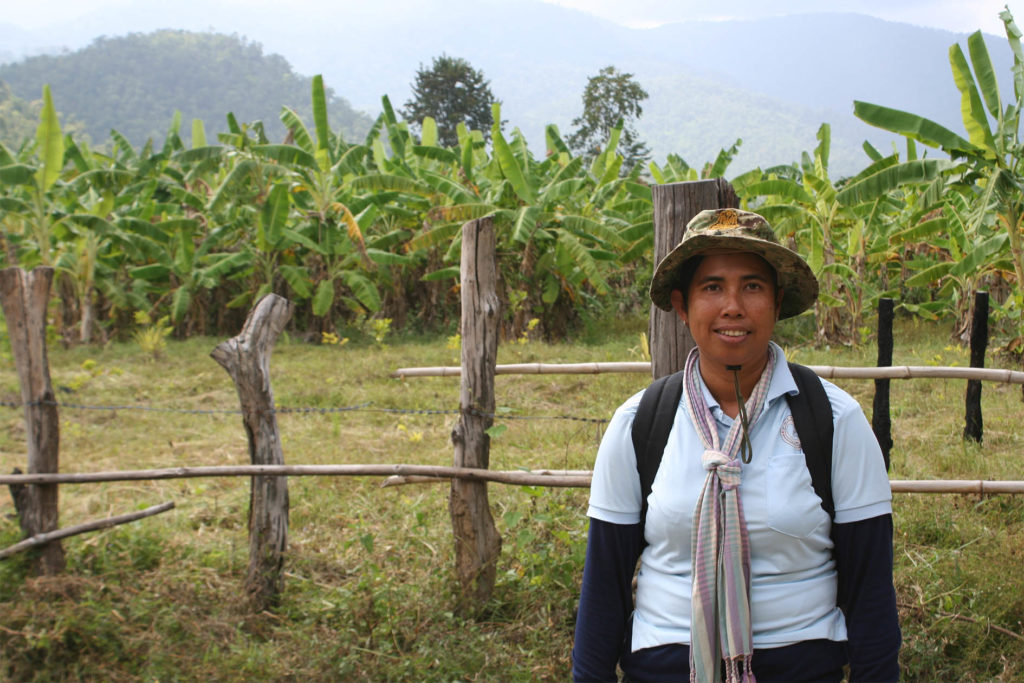 Savry, a volunteer malaria worker, smiles at the camera. Behind her, a fence, lush green forest and mountain range in the distance.