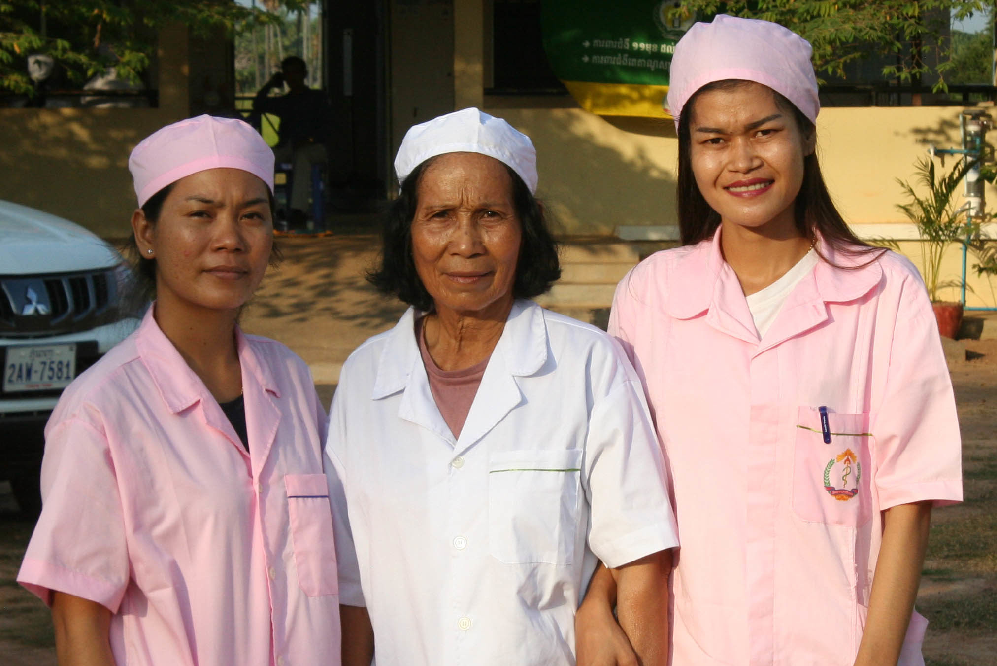 Three delivery room nurses stand arm in arm outside a clinic. The woman in the middle wears a white uniform and the women on either side of her wear pink. All are smiling.