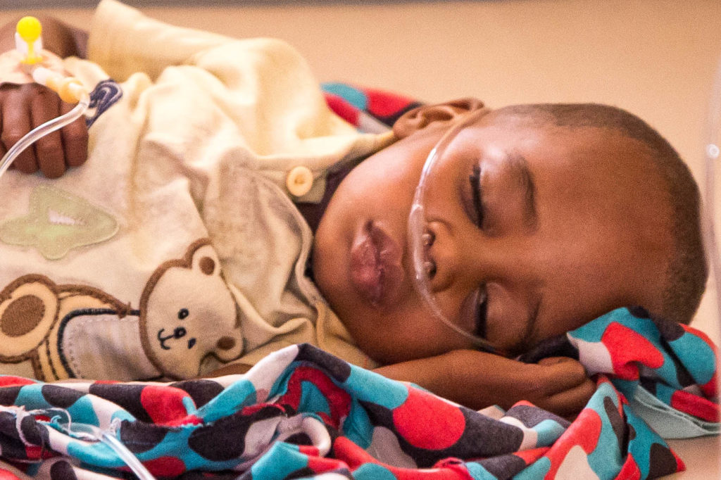 A toddler lies on a blue and red blanket with his eyes closed. He is receiving oxygen.