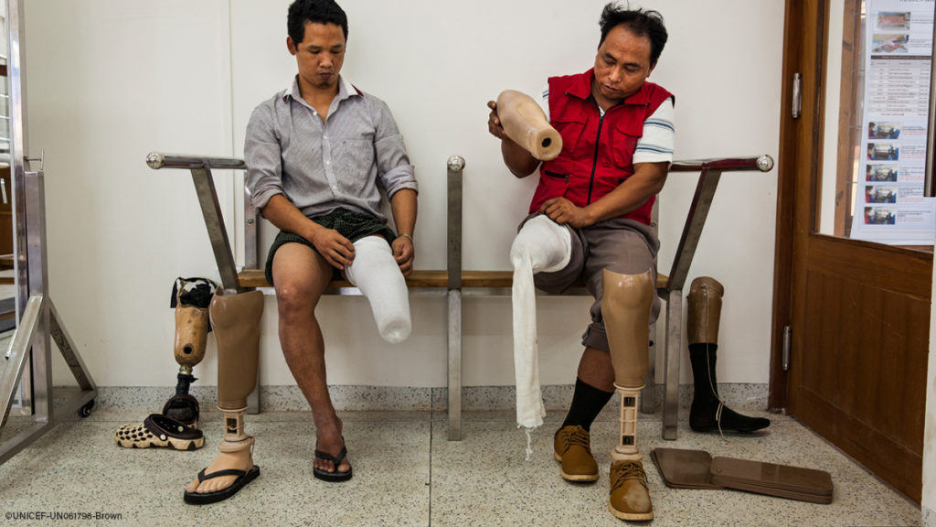 Two men on a bench are putting on prosthetic liners and socks with a prosthetic leg in front of each ready to be put on as well. Credit: UNICEF, Brown