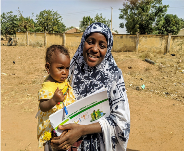 A woman stands outside in Katsina State, Nigeria. She smiles at the camera, holding her infant daughter in her arms.