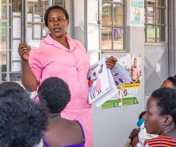 A nurse in pink uniform stands in front of a row of windows, giving a family planning talk to women as they wait to immunize their children.