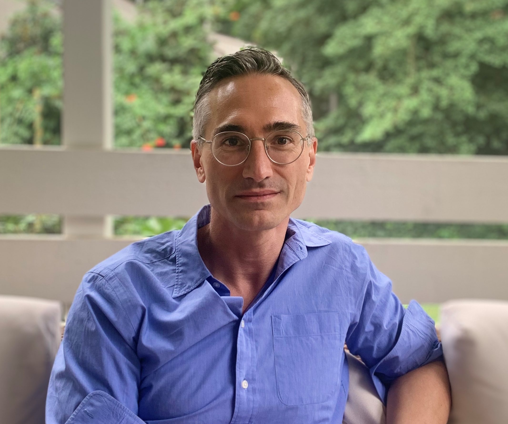 Arnaud Le Menach sits on a porch. He looks at the camera, wearing glasses and a blue dress shirt.