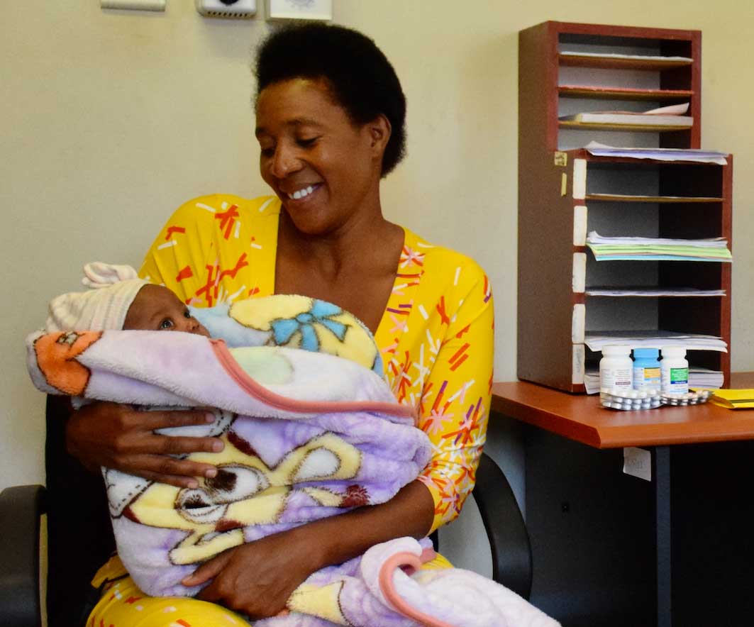 A mother in a yellow dress smiles down at her baby, who is bundled in blankets. She sits across from a doctor at a desk with a computer.