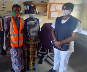 A SMAG in an orange vest stands with a patient and a nurse beside a clinic bed. They wear masks and look at the camera.