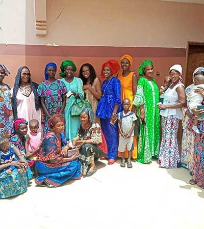 A group of brightly dressed mothers stand in a semi-circle. Some of the women hold their infants. They are smiling. The courtyard has terracotta walls and is filled with sun. 