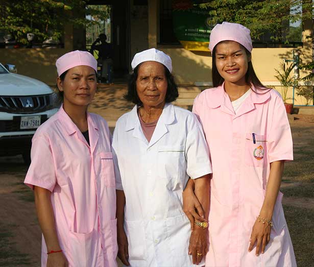 Three delivery room nurses stand arm in arm outside a clinic. The woman in the middle wears a white uniform and the women on either side of her wear pink. All are smiling.